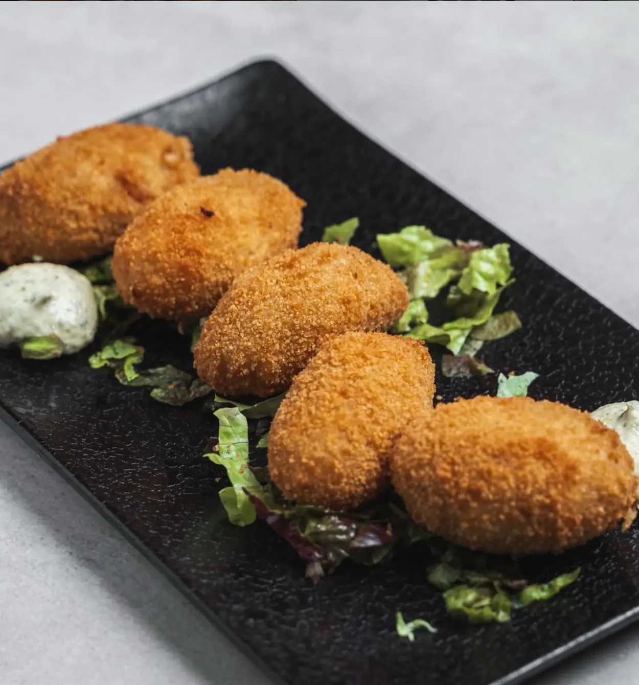 Cinco croquetas caseras doradas sobre lechuga en una bandeja negra, con salsa blanca a los lados