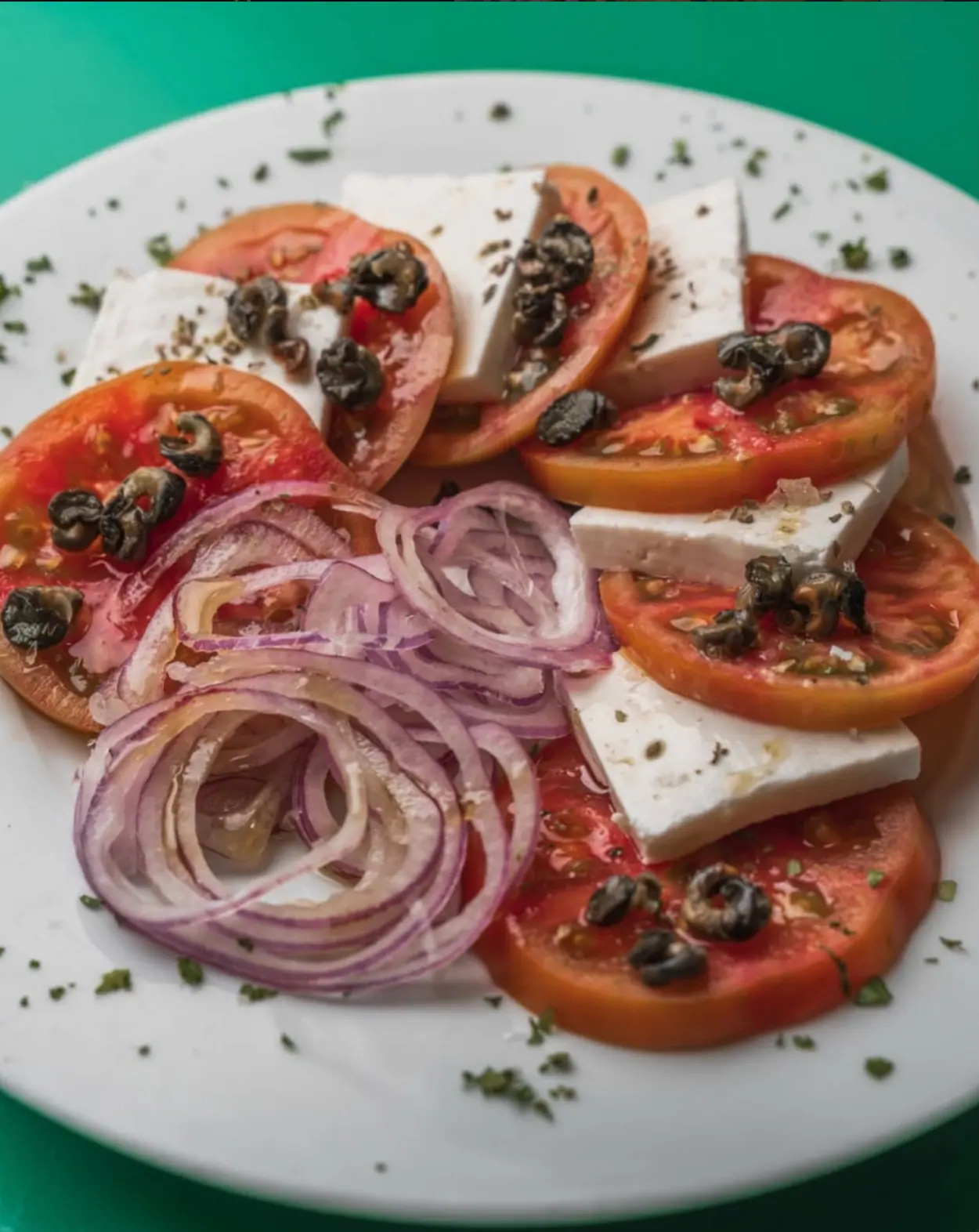 Ensalada de tomate en rodajas con queso blanco, cebolla morada y orégano sobre un mantel verde