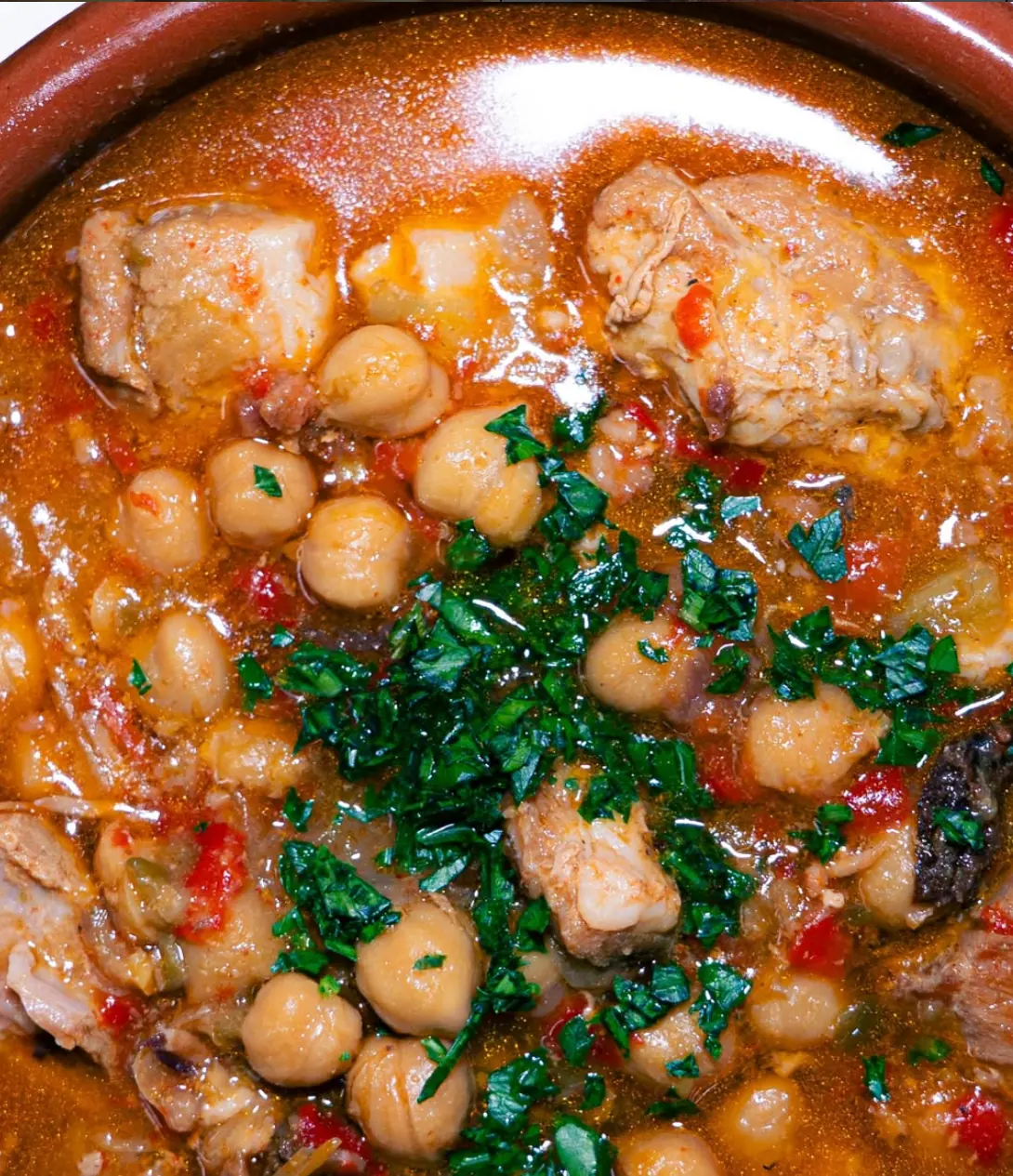 Cazuela de barro con garbanzos, carne y perejil picado en un caldo rojizo