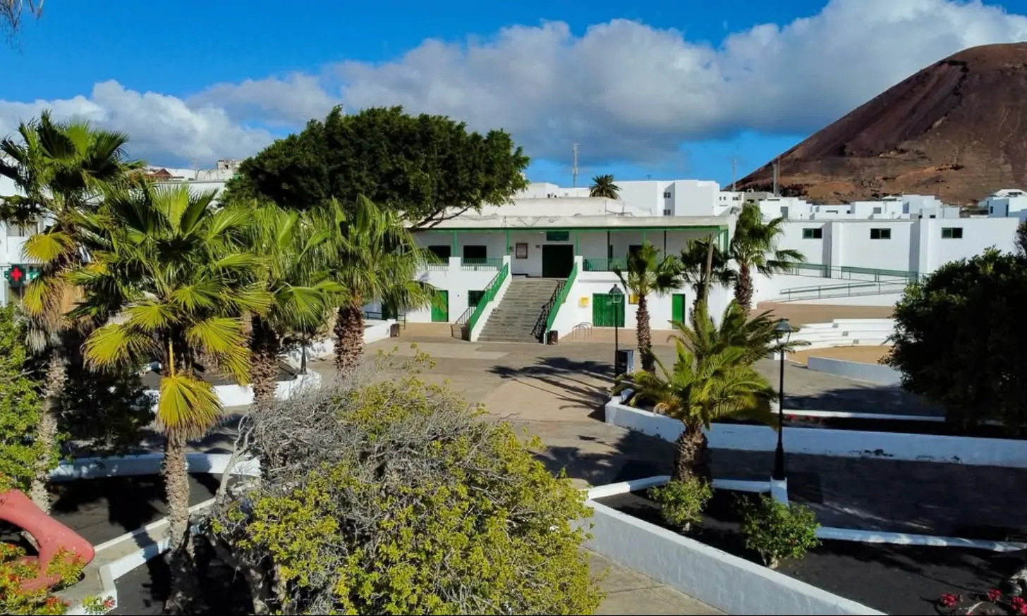 Fachada blanca del Teleclub de Tahíche, con escalinata, barandillas y puertas verdes, frente a la plaza con palmeras y el volcán al fondo