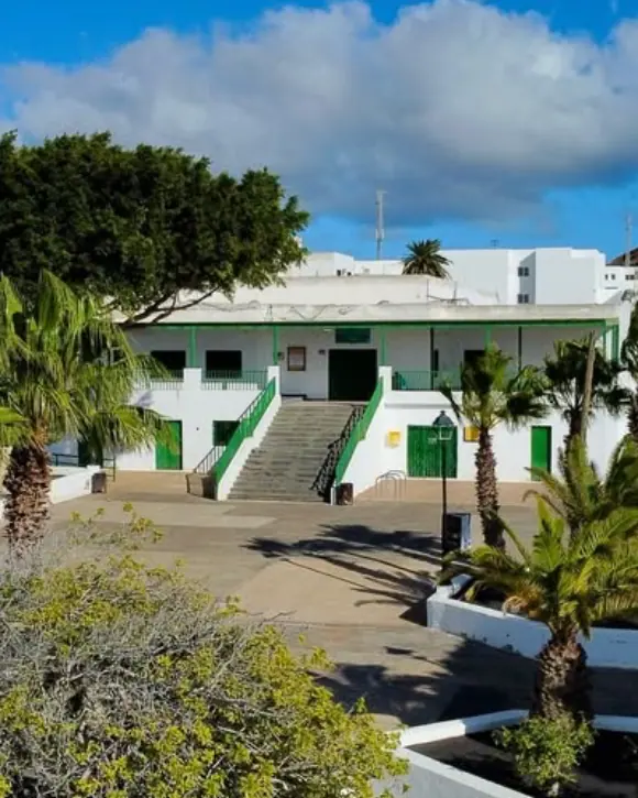 El Teleclub de Tahíche entre palmeras: casa blanca de puertas verdes con escalinata a la plaza