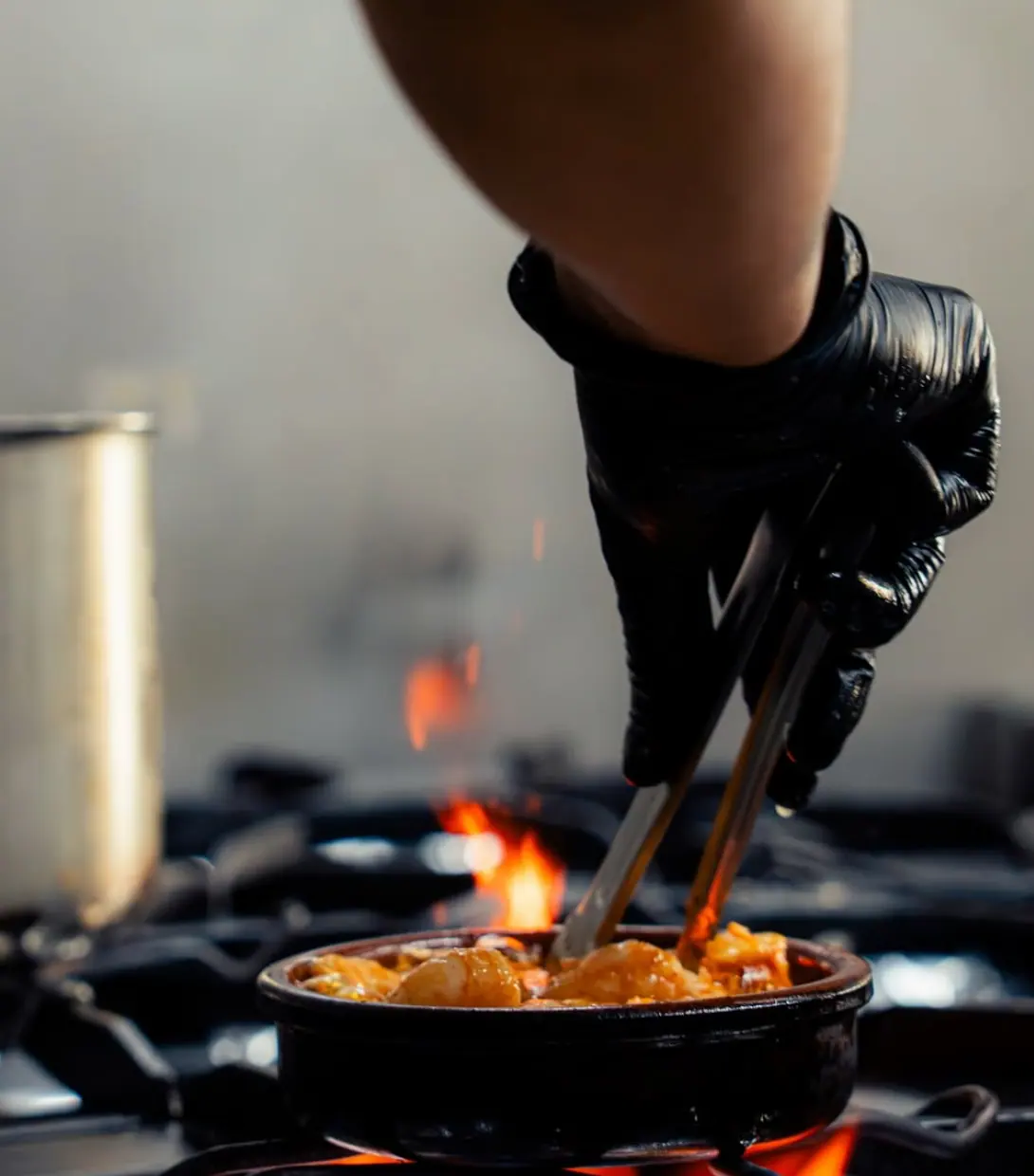 Mano con guante negro removiendo con pinzas una cazuela de barro sobre el fuego de la cocina