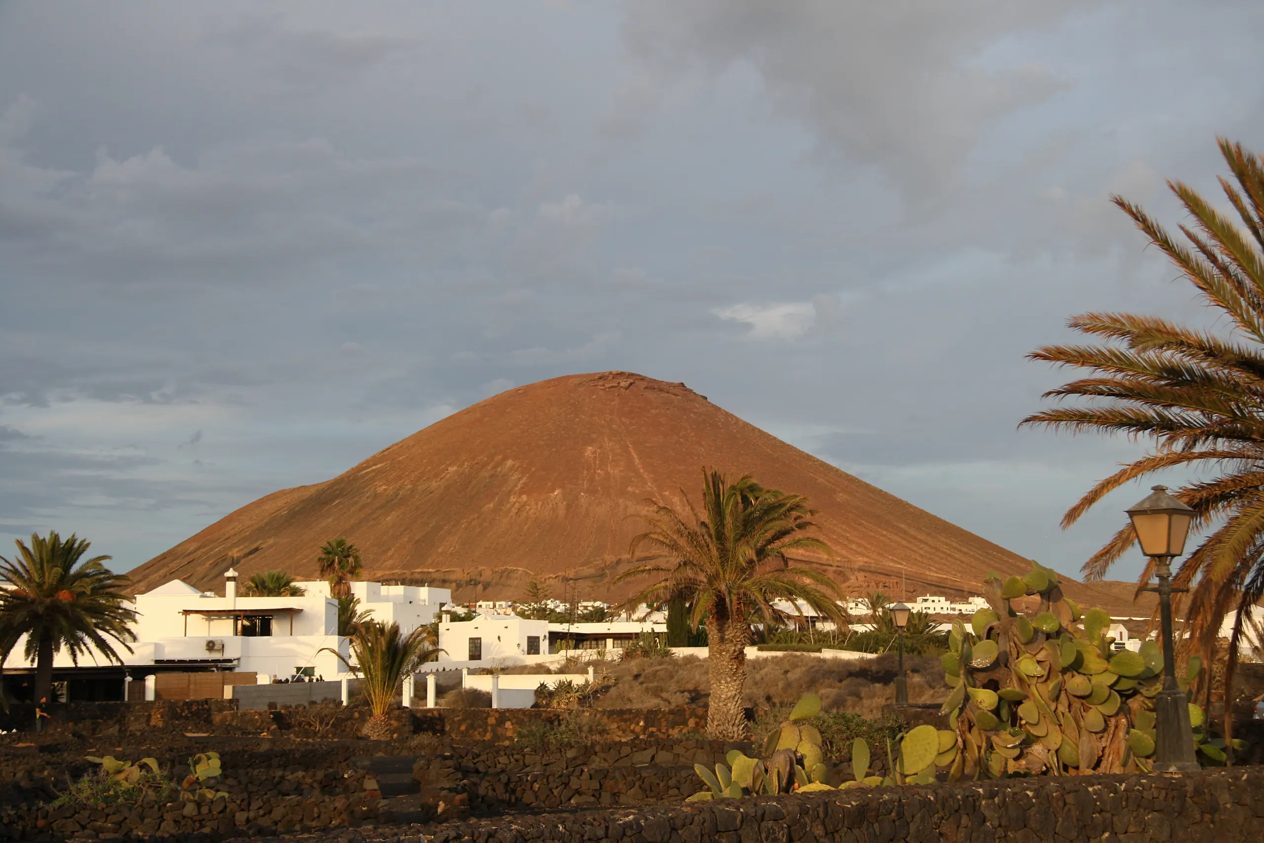 La montaña de Tahíche iluminada por el sol de la tarde, sobre casas blancas, palmeras y muros de piedra volcánica